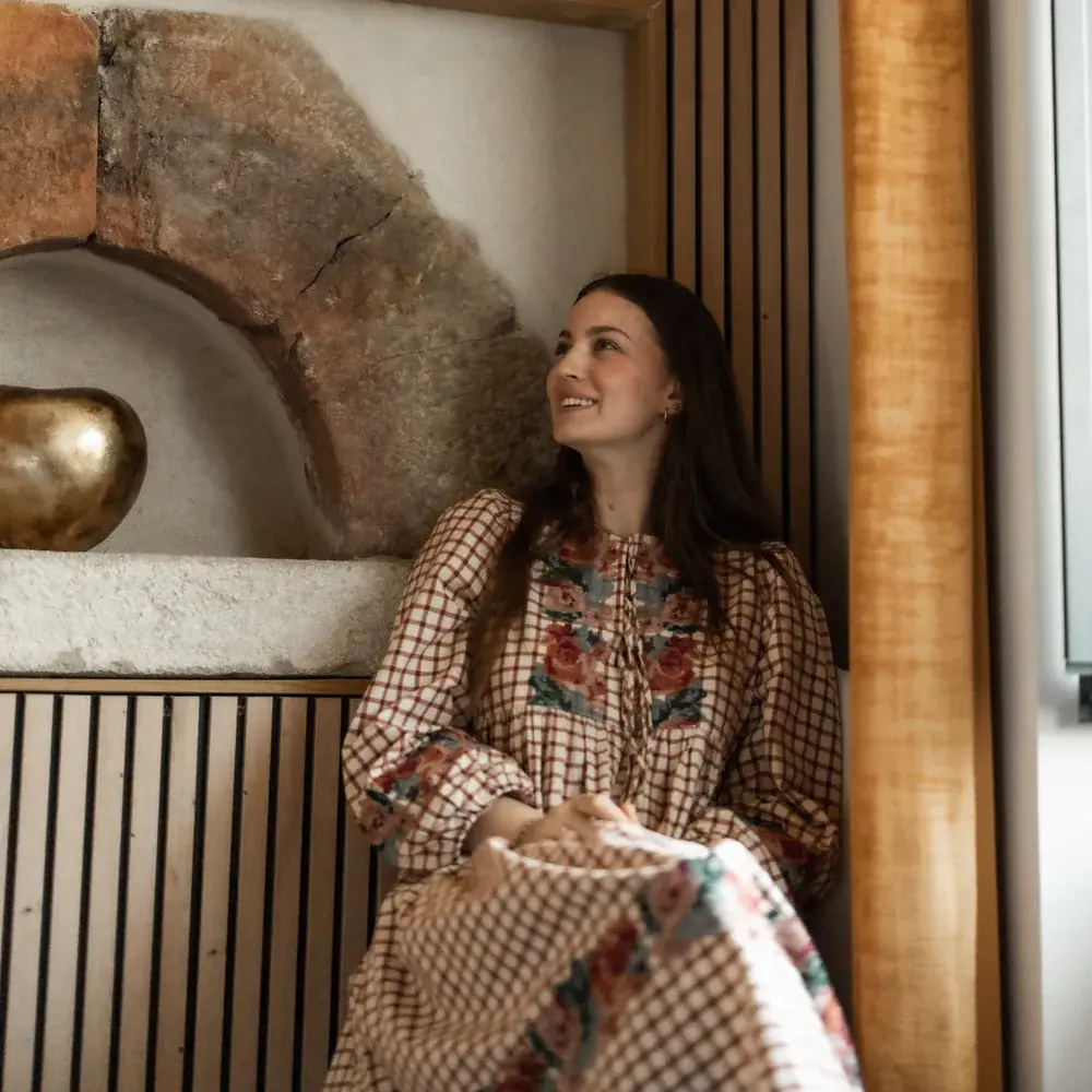Assise dans un intérieur en bois clair, robe fleurie, sourire aux lèvres.